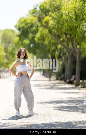 Une femme est debout sur un trottoir devant des arbres. Elle porte un débardeur blanc et un pantalon gris Banque D'Images