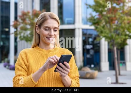 Une femme joyeuse dans un pull jaune utilisant joyeusement son smartphone tout en se tenant debout dans un paysage urbain moderne avec des arbres et des immeubles de bureaux en arrière-plan. Banque D'Images