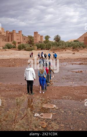 Les touristes traversent une rivière peu profonde jusqu'au village fortifié d'Aït Benhaddou sous un ciel couvant. Banque D'Images