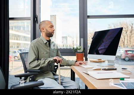 homme afro-américain handicapé atteint de myasthénie grave est assis à un bureau engrossé dans son travail, face à un écran d'ordinateur dans un cadre de bureau typique de co Banque D'Images