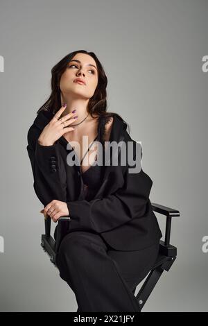 Femme élégante de taille plus profonde dans la pensée, menton reposant sur la main, assis élégamment dans une chaise sur un fond gris. Banque D'Images