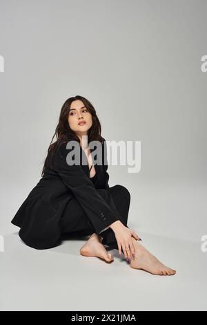 Une belle femme grande taille dans une tenue élégante s'assoit gracieusement avec ses jambes croisées sur un fond gris. Banque D'Images