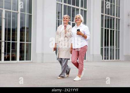 Joyeuses femmes âgées souriantes marchent et conversent en ville, profitant d'une promenade tranquille ensemble, habillées dans des vêtements élégants de fantaisie, mode féminin mature Banque D'Images