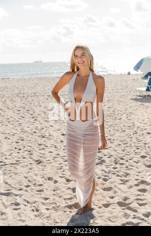 Une jeune et belle femme blonde se tient gracieusement au sommet d'une plage de sable à Miami, embrassant l'environnement serein. Banque D'Images