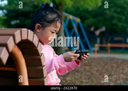 Un enfant utilisant ou jouant avec un smartphone à l'aire de jeux au lieu de jouer. Une petite fille regardant des vidéos sur un smartphone. Banque D'Images