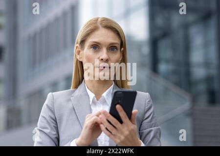 Une femme d'affaires confiante en costume gris utilise son smartphone dans un contexte urbain moderne, véhiculant un sens de la communication professionnelle et du professionnalisme dans un environnement urbain animé. Banque D'Images