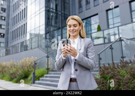 Une femme d'affaires en costume gris utilise son smartphone à l'extérieur des immeubles de bureaux modernes, reflétant un mélange de professionnalisme et de technologie dans un environnement d'entreprise. Banque D'Images