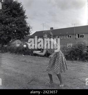 1964, historique, dehors dans un champ, derrière un lotissement, une jeune adolescente portant une robe d'été et des tongs, jouant à un jeu de chauve-souris et de balle, Angleterre, Royaume-Uni. Banque D'Images