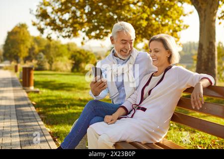 Heureux couple senior assis sur le banc dans le parc en utilisant le téléphone portable ensemble et rire à l'extérieur. Banque D'Images