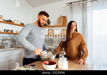 Un jeune couple marié beau en chandails cuisinent des biscuits de Noël ensemble dans la cuisine à la maison. Homme attrayant fouet les œufs pour la pâte avec un fouet. Banque D'Images