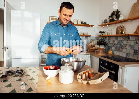 Un jeune homme beau cuisine dans la cuisine à la maison. Un homme attrayant casse des œufs dans un bol en métal. Assiettes, une casserole de sucre et un pot à biscuits sont sur le Banque D'Images