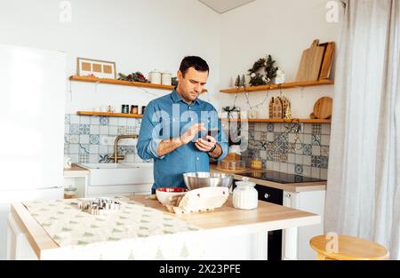 Un homme attrayant dans une chemise en denim cuisine dans la cuisine à la maison. Un jeune homme tient un téléphone et lit une recette. Un mâle fait des biscuits en pain d'épices Banque D'Images