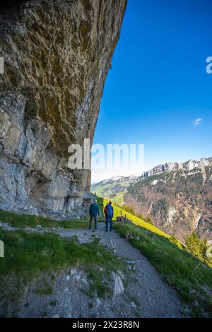 Description : les touristes sur le sentier alpin à la cabane Äscher se tiennent en dessous et regardent l'énorme mur de roche surplombant d'en bas. Seealpsee, Appenzell, Suisse, Banque D'Images