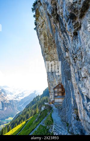 Description : cabane suisse en bois Gasthaus Aescher-Wildkirchli en dessous d'un mur de roche massif et d'une chaîne de montagnes en arrière-plan. Seealpsee, Appenze Banque D'Images