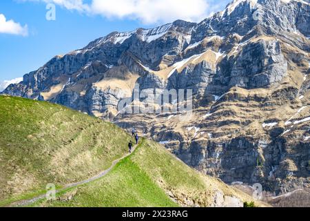 Description : les touristes marchent le long d'un sentier de randonnée sur une prairie alpine suisse verte avec une énorme paroi rocheuse escarpée en arrière-plan par une journée ensoleillée. Seealpse Banque D'Images