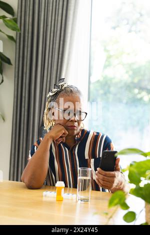 Femme aînée afro-américaine portant des lunettes regardant un smartphone à la maison et prenant des médicaments Banque D'Images