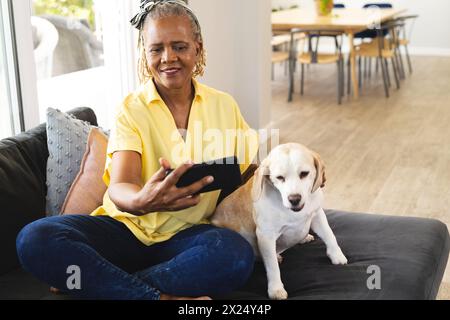 Femme aînée afro-américaine assise avec un chien à la maison, tenant un smartphone Banque D'Images