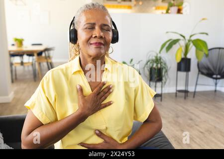 Une femme afro-américaine senior appréciant la musique avec des écouteurs à la maison, la main sur le cœur, l'espace de copie Banque D'Images