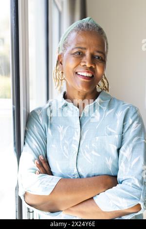 Femme aînée afro-américaine debout près de la fenêtre à la maison, les bras croisés, souriante Banque D'Images