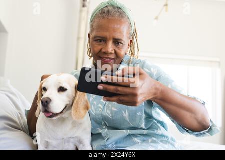 Femme aînée afro-américaine tenant un smartphone, prenant selfie avec un chien à la maison Banque D'Images