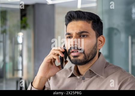 Un Indien semble tendu et frustré tout en parlant sur son téléphone portable dans un bureau moderne, montrant des signes de colère et de stress. Banque D'Images