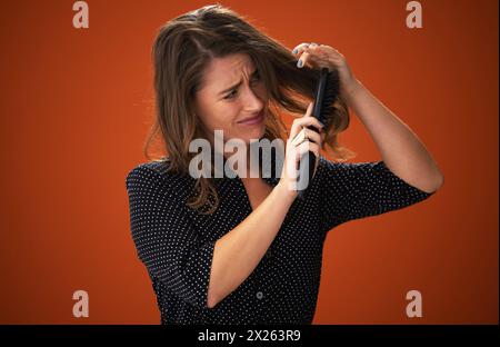 Frustré, femme et brosse pour les cheveux, sec et emmêlé d'humidité, enchevêtrement et difficile à peigner. Fond rouge, texture et douleur des dommages de Banque D'Images