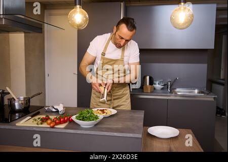 Bel homme caucasien, un chef masculin dans un tablier beige, râpant du fromage parmesan dans un plat de spaghettis avec de la sauce tomate, cuisinant le dîner selon Banque D'Images