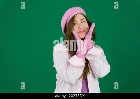 Jeune fille femme avec un béret rose sur sa tête mode France sur un fond vert chromakey. différents gants tricotés d'automne rose hiver sur les mains. Jolie fille portant un chapeau français rose. Banque D'Images