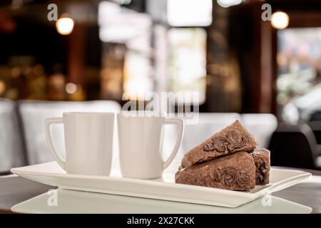 Trois morceaux de gâteau gaufre au chocolat avec deux tasses sur une plaque en céramique blanche sur la table, fond de café. Banque D'Images