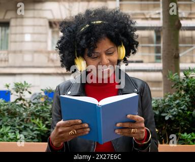 Femme afro-américaine d'âge moyen lisant un livre assis sur un banc dans une ville Banque D'Images