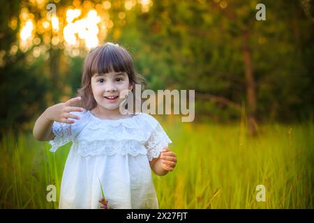 Une charmante petite fille heureuse dans une robe blanche se tient dans un champ près d'une forêt de conifères en été Banque D'Images