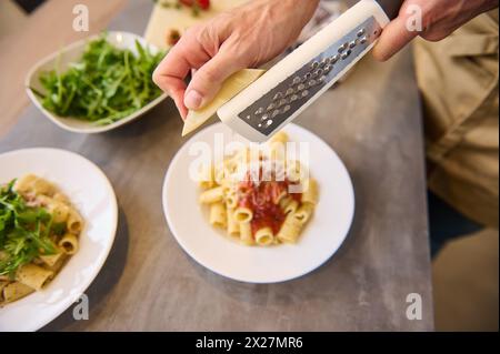 Directement au-dessus des mains du chef à l'aide d'une râpe, râper le fromage sur des pâtes fraîchement cuites avec de la sauce tomate, assaisonner et étaler le repas avant de le servir. Banque D'Images