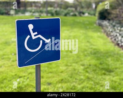 Un petit panneau bleu pour handicapés en fauteuil roulant se dresse contre un fond de jardin flou, symbolisant l'accessibilité et l'inclusivité dans les espaces extérieurs. Banque D'Images