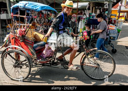 Samut Sakhon, Thaïlande – 18 février 2023 : devant des stands de fruits et de boissons, un chauffeur de taxi tricycle donne une pancarte avant de descendre de son tricycle. Banque D'Images