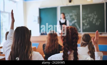 Groupe intelligent de jeunes écoliers levant tous les mains en l'air pour répondre à une question posée par l'enseignante, vue de derrière Banque D'Images