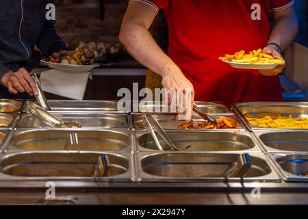 Deux hommes se servent de nourriture et de frites avec des pinces dans leurs mains à une table avec de la nourriture servie dans des seaux en acier inoxydable à un amateur de nourriture d'un restaurant Banque D'Images