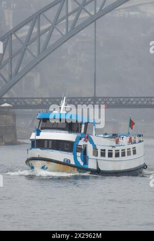 Ferry touristique au premier plan naviguant le long du fleuve Douro en passant sous le pont en acier Don Luis I à Porto sur une journée très brumeuse et pluvieuse avec tr Banque D'Images
