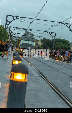 Vue de face du métro de Porto circulant sur les pistes du pont Dom Luis au crépuscule, avec les lanternes au sol allumées avec la lumière orange et les touristes Banque D'Images