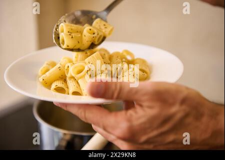 Gros plan verser des pâtes italiennes sur une assiette blanche avant de les plaquer et de les servir aux clients. Le chef tient une assiette avec des pâtes italiennes fraîchement cuites Banque D'Images