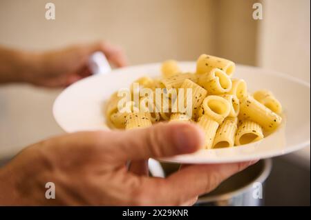 Gros plan verser des pâtes italiennes sur une assiette blanche avant de les plaquer et de les servir aux clients. Le chef tient une assiette avec des pâtes italiennes fraîchement cuites. Nourriture Banque D'Images