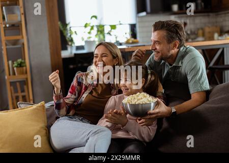 Une famille de trois est confortablement nichée sur un canapé, leurs visages reflétant l'excitation et l'attention comme ils partagent un bol de pop-corn pendant un susp Banque D'Images