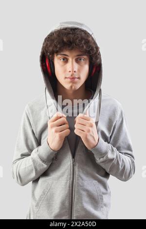 Beau jeune homme porte des écouteurs et un sweat-shirt à capuche et est isolé sur un fond gris. Il regarde dans la caméra avec ses yeux bleus whi Banque D'Images