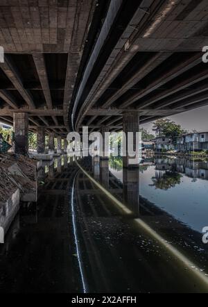 Bangkok, Thaïlande - avril 20, 2024 - vue en perspective des piliers en béton sous Khlong Toei Expressway avec réflexion calme de l'eau de surface et les maisons sont Banque D'Images