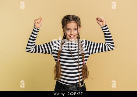 Je suis fort et en bonne santé. Jeune fille mignonne montrant des biceps et regardant confiant ressentant la force de puissance pour se battre pour les droits, l'énergie pour gagner la victoire de succès. Enfant de preteen 10 ans sur fond beige Banque D'Images