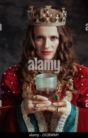 reine médiévale en robe rouge avec gobelet et couronne sur fond gris foncé. Banque D'Images