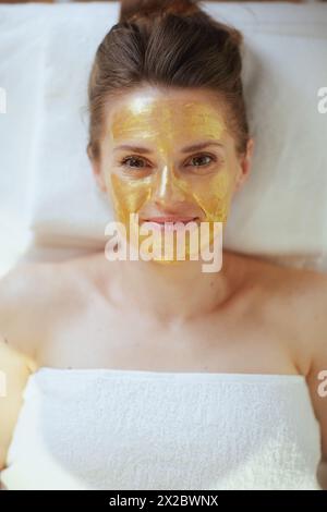 Temps de soins. Vue supérieure de la femme moderne détendue dans le salon de spa avec masque cosmétique doré sur le visage couché sur la table de massage. Banque D'Images