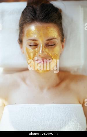 Temps de soins. Vue supérieure de la femme moderne d'âge moyen détendue dans le salon de spa avec masque cosmétique doré sur le visage couché sur la table de massage. Banque D'Images