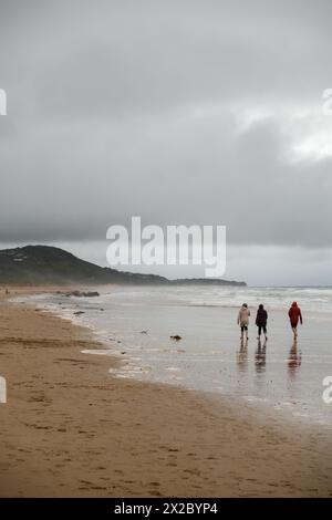 Une scène sereine de trois individus marchant tranquillement le long du rivage, du sable humide sous leurs pieds, sous un ciel nuageux avec des collines ajoutant une toile de fond. Banque D'Images