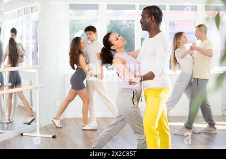 Jeune femme souriante portant des vêtements de sport dansant dans la danse de rumba de couple avec partenaire afro-américain pendant la classe de groupe au salon de danse Banque D'Images