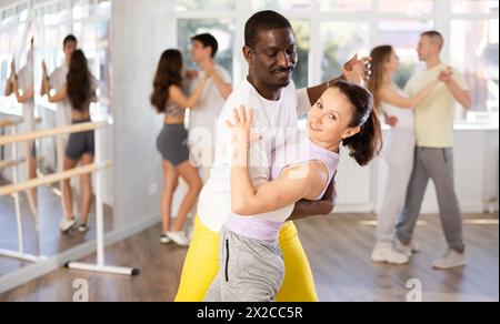 Jeune femme souriante portant des vêtements de sport dansant dans la danse samba couple avec partenaire afro-américain pendant la classe de groupe au salon de danse Banque D'Images